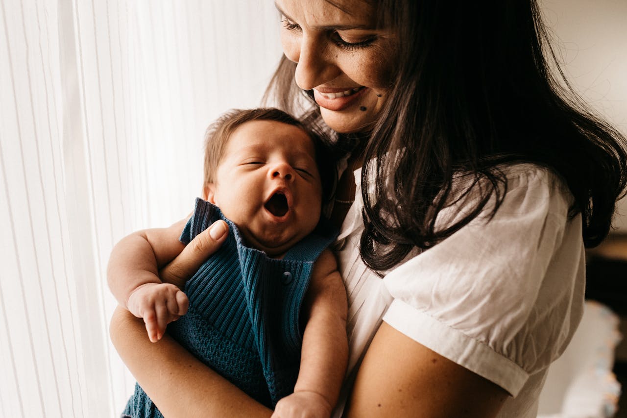 A loving mother smiles while holding her yawning newborn baby near a sunny window.